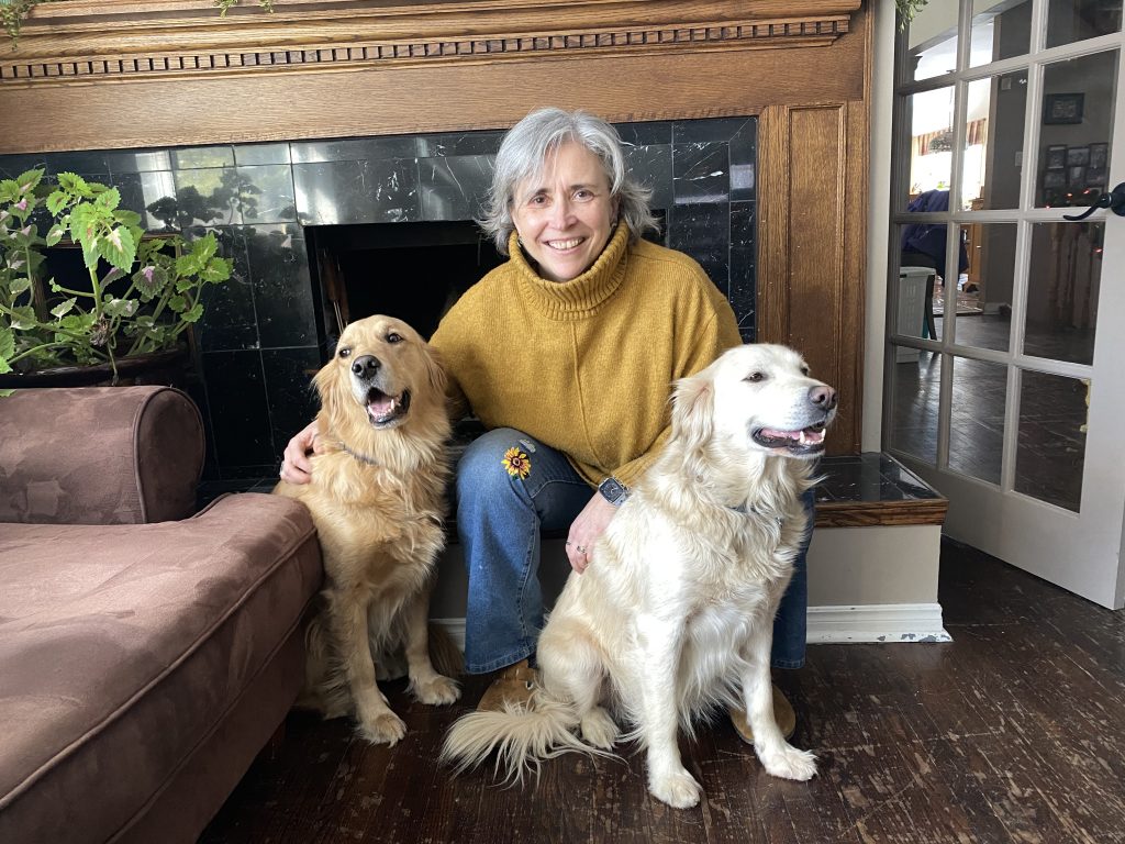 Martine in a yellow sweater crouching with her two golden retrievers, Alice and Penny.